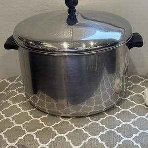May include: A large, cylindrical stainless steel stockpot with a polished finish. It features two black side handles and a matching lid with a central black knob. The pot has a wide, flat base and straight vertical walls, providing a deep cooking capacity.