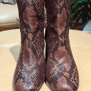 May include: A pair of brown and black snakeskin-patterned ankle boots. The boots have a medium heel and a textured surface. The pattern is a mix of dark brown and black, creating a reptile-like appearance. The boots are standing upright.