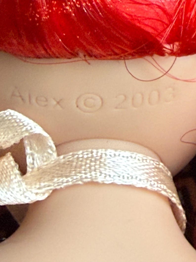 May include: Close-up of a doll's head with bright red hair. The doll's neck is visible, with a cream-colored ribbon tied around it. The name "Alex" and the year "2003" are imprinted on the doll's neck.