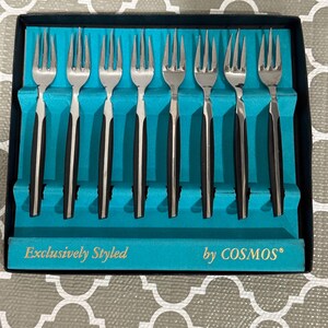 May include: A set of eight forks with stainless steel tines and black handles, presented in a teal box. The box is labeled "Exclusively Styled by COSMOS®". The forks are arranged neatly in individual slots.