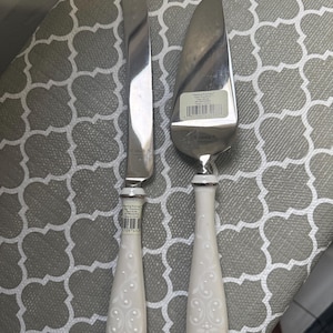 May include: A silver-toned cake knife and server set featuring white ceramic handles with a raised floral scroll pattern. Each piece is marked with the Lenox brand name and includes a product label with a barcode and the text Wedding Promises.