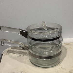 Op de afbeelding: Transparante glazen kookpot met een deksel en twee transparante glazen handvatten. De pot heeft een metalen band in het midden. Ontworpen om te koken, staat hij op een wit oppervlak.