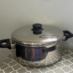 May include: A polished stainless steel stockpot with a matching lid. The pot features two black heat-resistant side handles and a central black knob on the lid. The reflective metal surface shows a clear, smooth finish throughout the exterior.