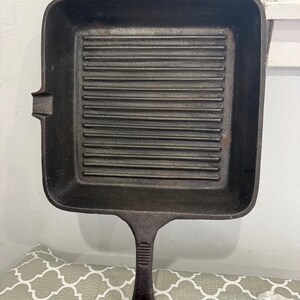 May include: A square cast iron grill pan with a dark, textured finish. The pan features a series of parallel raised ridges across the cooking surface, a long handle with a grooved grip, a small pouring spout on the side, and a top hanging loop.