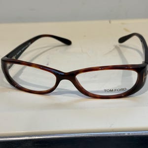 以下が含まれることがあります： べっ甲柄のフレームが特徴的な、茶色と黒のトムフォードの眼鏡。丸みを帯びた形状で、控えめなキャットアイデザイン。レンズにはトムフォードのロゴが見えます。