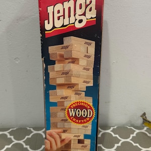 Könnte beinhalten: Eine Jenga-Spielbox mit dem Wort "Jenga" in Weiß auf rotem Hintergrund. Das Spiel besteht aus präzisionsgefertigten Holzblöcken. Die Blöcke sind zu einem Turm gestapelt, wobei eine Hand einen Block entfernt.
