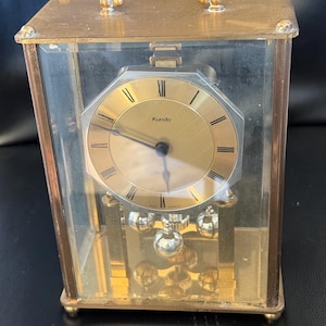 May include: A vintage brass and glass clock with the brand name "Kundo" on the gold-colored clock face. The clock has a handle on top and a pendulum with multiple silver spheres. The clock is encased in a rectangular glass box.