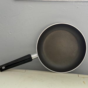 May include: A non-stick frying pan with a black handle and a silver rim. The pan is round and has a dark interior. The handle is made of black plastic and is attached to the pan with a silver metal piece.