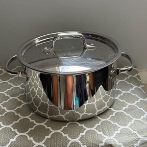 May include: A stainless steel stockpot with a matching lid. The pot has two side handles and a large handle on the lid. The pot is sitting on a gray and white patterned surface. The pot is reflective.