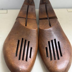 May include: Pair of wooden shoe trees with a rich brown finish. Each tree features a rounded knob at the top and a series of vertical slots near the toe. The shoe trees are designed to maintain the shape of shoes and absorb moisture.