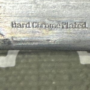 K&ouml;nnte beinhalten: Nahaufnahme eines metallischen Objekts mit der Pr&auml;gung "Hard Chrome Plated". Die Oberfl&auml;che hat eine gl&auml;nzende, reflektierende Qualit&auml;t, die auf eine Chromoberfl&auml;che hindeutet. Der Hintergrund ist strukturiert und gr&uuml;n.
