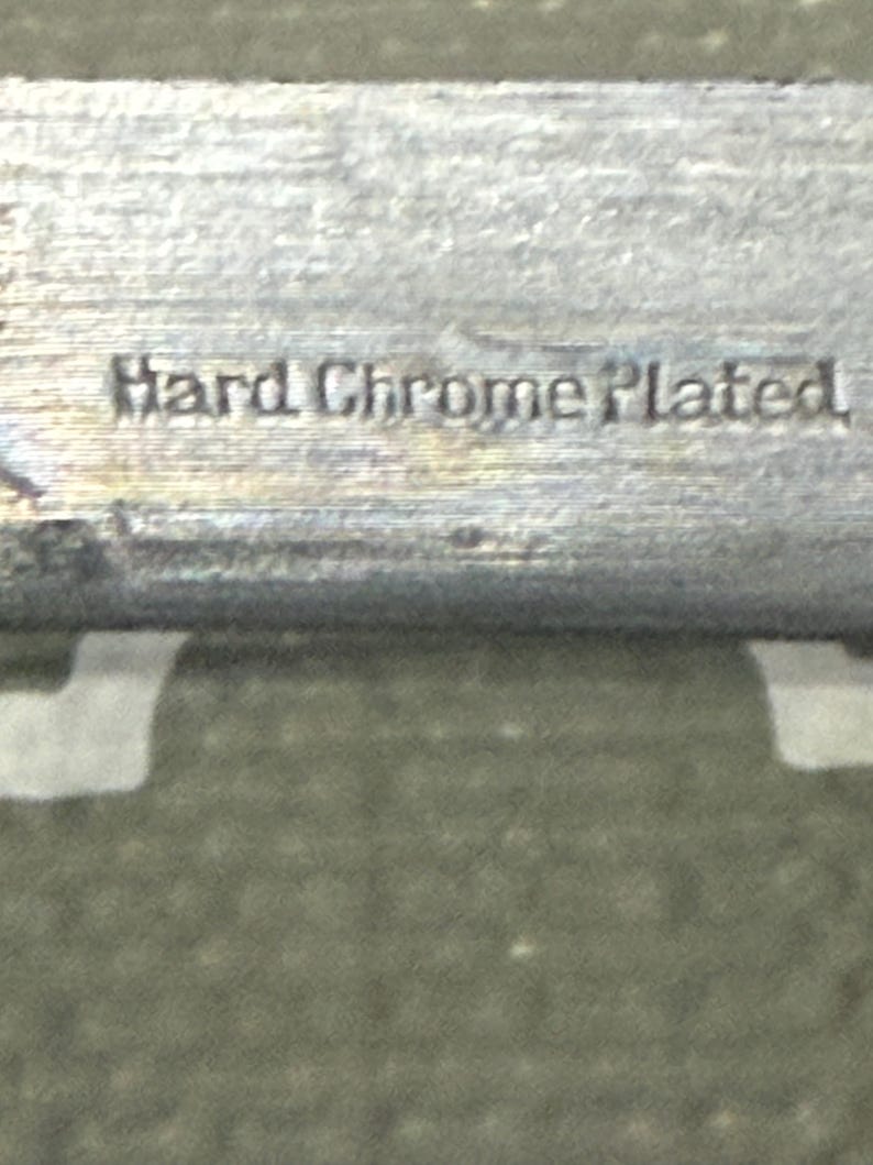 K&ouml;nnte beinhalten: Nahaufnahme eines metallischen Objekts mit der Pr&auml;gung "Hard Chrome Plated". Die Oberfl&auml;che hat eine gl&auml;nzende, reflektierende Qualit&auml;t, die auf eine Chromoberfl&auml;che hindeutet. Der Hintergrund ist strukturiert und gr&uuml;n.