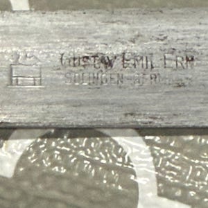 K&ouml;nnte beinhalten: Nahaufnahme eines silbernen Metallobjekts mit dem Text "GUSTAV EMIL ERN SOLINGEN GERMANY" auf der Oberfl&auml;che. Das Objekt hat ein kleines, stilisiertes Geb&auml;udesymbol.