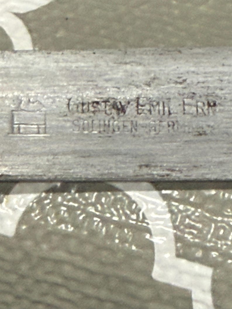 K&ouml;nnte beinhalten: Nahaufnahme eines silbernen Metallobjekts mit dem Text "GUSTAV EMIL ERN SOLINGEN GERMANY" auf der Oberfl&auml;che. Das Objekt hat ein kleines, stilisiertes Geb&auml;udesymbol.