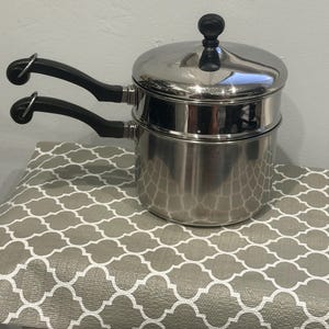 Può includere: Un set di pentole a bagnomaria in acciaio inossidabile con finitura lucida, due manici neri resistenti al calore con anelli di sospensione in metallo e un coperchio abbinato con pomello nero. Il design include una pentola profonda e un inserto.