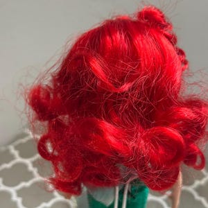 May include: A doll with vibrant red, curly hair. The doll's hair is styled in loose curls and waves. The doll is wearing a green outfit. The background is a light gray color.