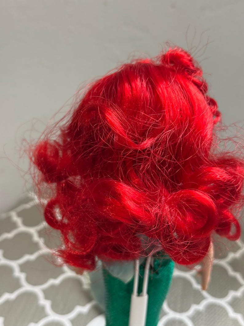 May include: A doll with vibrant red, curly hair. The doll's hair is styled in loose curls and waves. The doll is wearing a green outfit. The background is a light gray color.