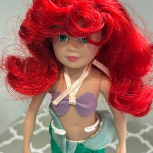 May include: A doll of a mermaid with bright red hair, blue eyes, and a purple seashell top. The doll has a green sequined tail and a light blue and white collar.