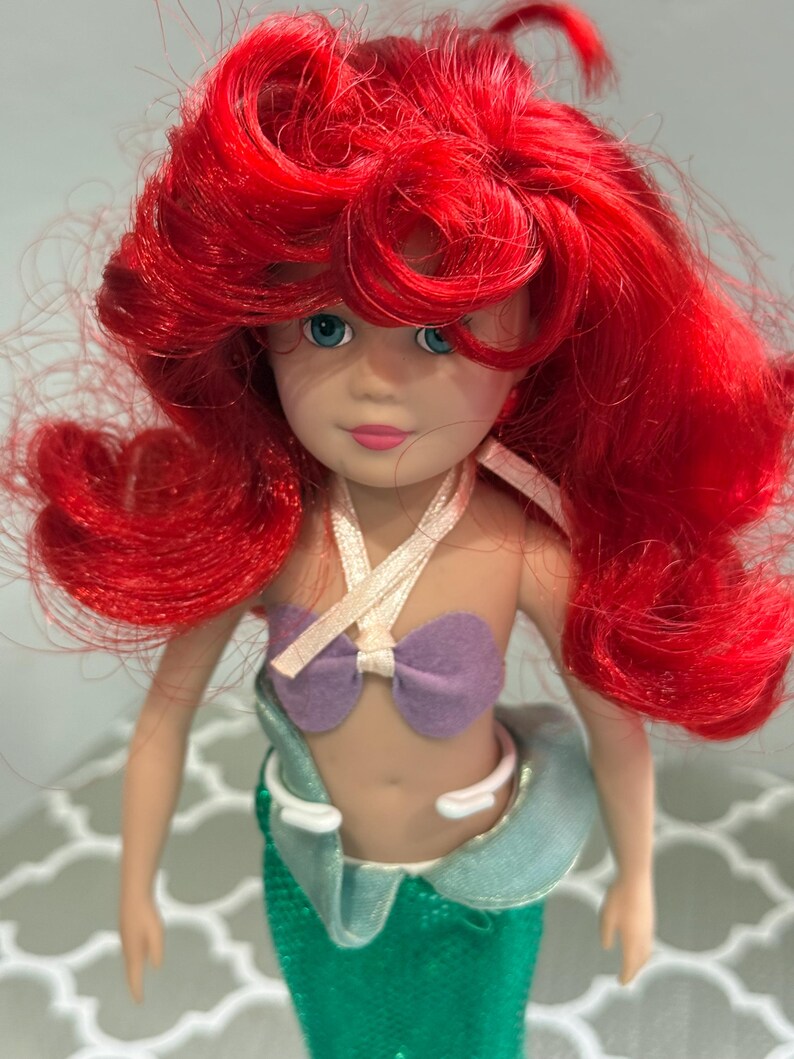 May include: A doll of a mermaid with bright red hair, blue eyes, and a purple seashell top. The doll has a green sequined tail and a light blue and white collar.