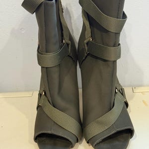 Könnte beinhalten: Ein Paar olivgrüne Stiefeletten mit offener Spitze. Die Stiefel haben mehrere olivgrüne Riemen mit goldfarbener Hardware. Die Absätze sind hoch und die Stiefel haben ein schlankes, modernes Design. Die Stiefel sind aus einem weichen, matten Material.