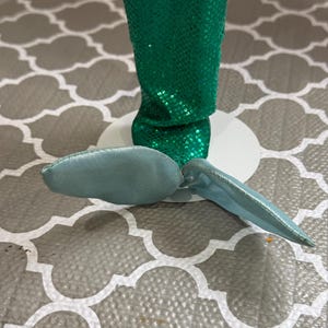 May include: A doll with a green sequined mermaid tail and a light blue fin. The doll is standing on a white base. The background is a gray and white patterned surface.
