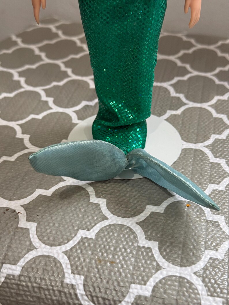 May include: A doll with a green sequined mermaid tail and a light blue fin. The doll is standing on a white base. The background is a gray and white patterned surface.