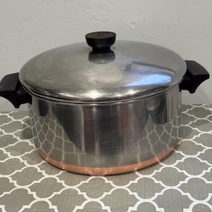Op de afbeelding: Een roestvrijstalen kookpan met bijpassend deksel, voorzien van zwarte kunststof handgrepen aan de zijkanten en een zwarte knop op het deksel. De onderkant van de pan heeft een koperkleurige rand en het buitenoppervlak is gepolijst.