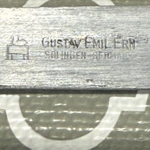 K&ouml;nnte beinhalten: Nahaufnahme eines Metallobjekts mit dem Text "GUSTAV EMIL ERN" und "SOLINGEN-GERMANY" auf der Oberfl&auml;che. Das Objekt hat ein strukturiertes, geb&uuml;rstetes Metallaussehen und ein kleines Logo.