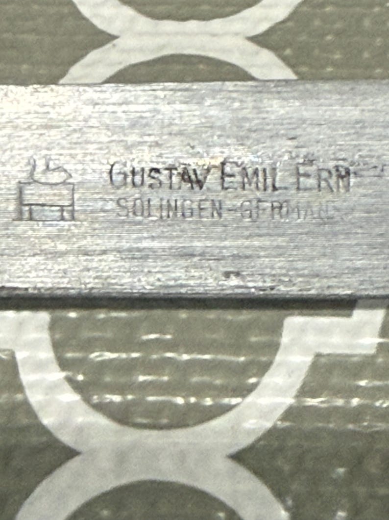 K&ouml;nnte beinhalten: Nahaufnahme eines Metallobjekts mit dem Text "GUSTAV EMIL ERN" und "SOLINGEN-GERMANY" auf der Oberfl&auml;che. Das Objekt hat ein strukturiertes, geb&uuml;rstetes Metallaussehen und ein kleines Logo.