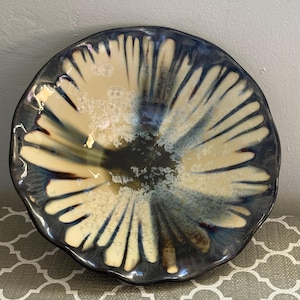 May include: A round ceramic bowl with a wavy, uneven rim. The interior features a crystalline glaze pattern with a dark, deep blue center that radiates outward into creamy white, petal-like streaks, creating a high-contrast, glossy finish.