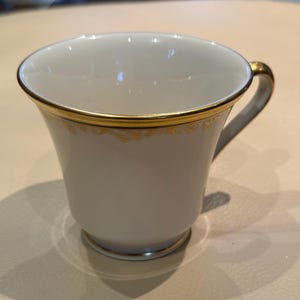 May include: White teacup with a gold rim and handle. The cup has a simple, elegant design with a delicate gold pattern along the rim. The cup is empty and appears to be made of porcelain.