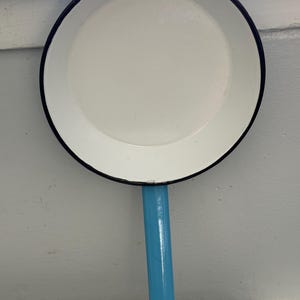 以下が含まれることがあります： 黒い縁と水色の持ち手が付いた白いエナメルフライパン。フライパンは丸く、シンプルなデザインでヴィンテージのようです。持ち手は長く細く、壁に掛かっています。