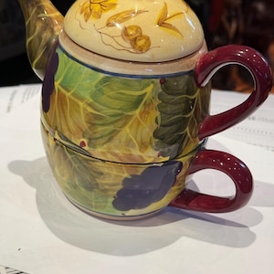 May include: A decorative ceramic teapot and teacup set. The teapot has a yellow lid with floral designs and a brown knob. The body is painted with green leaves and purple grapes. The teacup has a burgundy handle.