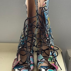 May include: Open-toe lace-up boots with a vibrant snakeskin print in shades of blue, pink, yellow, and black. The boots have black laces and a high-cut design. The background is a plain, neutral colour. The heels are not visible.