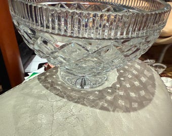May include: A clear, cut-glass bowl with a wide, ribbed rim and diamond-patterned body. The bowl sits on a short, decorative pedestal base. The glass reflects light, creating a sparkling effect. Ideal for serving fruit or as a decorative centerpiece.
