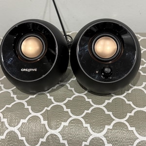 May include: A pair of spherical black computer speakers with metallic gold-colored center cones. The left speaker features the white text CREATIVE, while the right speaker includes a small black volume control knob and a tiny indicator light.