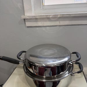 Peut inclure: Ensemble de batterie de cuisine en acier inoxydable comprenant une grande casserole, une petite casserole et un couvercle. Les casseroles ont des poignées noires et sont empilées. L'ensemble est posé sur une surface crème.
