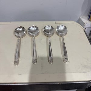 Può includere: Quattro cucchiai da zuppa d'argento con manici decorativi. I cucchiai sono disposti in fila, riflettendo la luce. I manici hanno un design ornato alla base. I cucchiai sono su una superficie chiara.