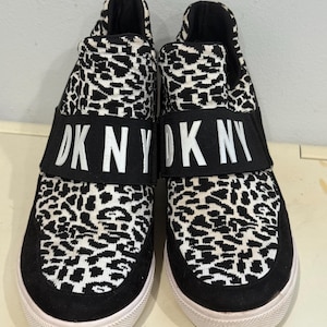 May include: Pair of slip-on sneakers with a black and white leopard print design. The shoes have a black band across the top with the text "DKNY" in white. The soles are white.