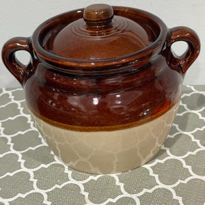 May include: A two-handled ceramic bean pot with a glossy dark brown glazed upper section and a smooth beige lower section. The pot features a matching removable lid with a central knob handle and two curved side handles for easy lifting.