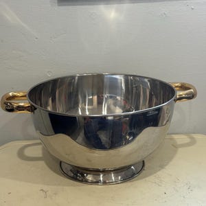 Puede incluir: Un cuenco grande y plateado pulido con dos asas doradas. El cuenco tiene un diseño amplio y abierto y se asienta sobre una base pequeña y redonda. El interior refleja el entorno. Las asas son curvas y añaden un toque decorativo.