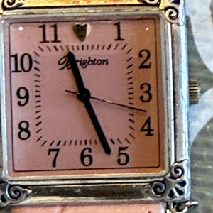 May include: A square-faced watch with a pink dial and a silver-colored frame. The watch face features black numerals, a heart at the 12 o'clock position, and the brand name "Brighton". The watch has a pink leather band.