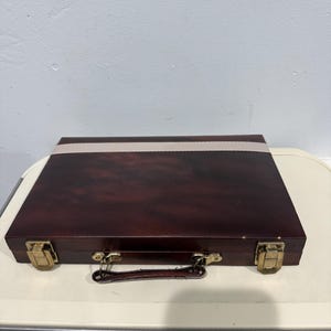 Puede incluir: Un maletín de madera marrón oscuro con herrajes de color latón. El maletín tiene una banda de color claro en la parte superior y un asa. El estuche está cerrado y parece estar vacío.