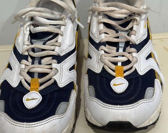 May include: Pair of white, navy blue, and yellow athletic shoes with white laces. The shoes have a mesh and leather upper with a visible Nike swoosh logo. The soles are black and gray.