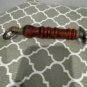 K&ouml;nnte beinhalten: Ein Vintage-Flaschen&ouml;ffner mit einem dunklen Holzgriff und Messingakzenten. Der Griff ist zylindrisch mit einem Ring an einem Ende und einem Metall&ouml;ffner am anderen. Zum Entfernen von Flaschenverschl&uuml;ssen.