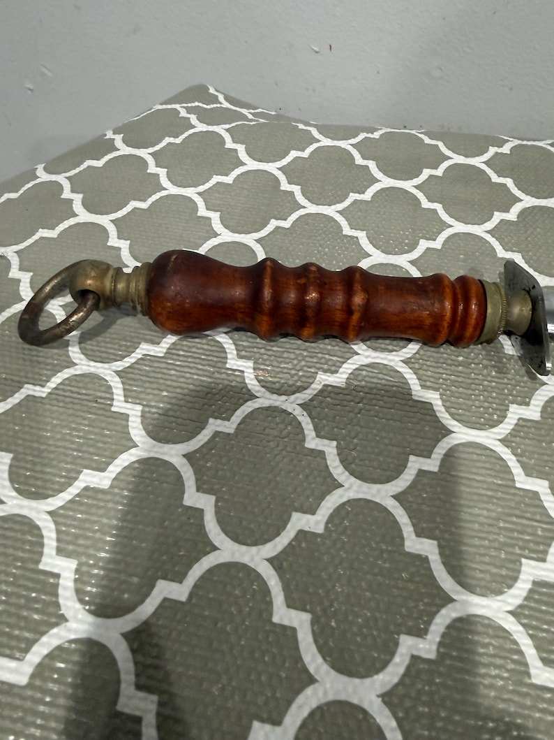 K&ouml;nnte beinhalten: Ein Vintage-Flaschen&ouml;ffner mit einem dunklen Holzgriff und Messingakzenten. Der Griff ist zylindrisch mit einem Ring an einem Ende und einem Metall&ouml;ffner am anderen. Zum Entfernen von Flaschenverschl&uuml;ssen.