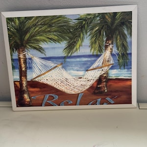 May include: A framed painting depicts a beach scene with two palm trees and a white hammock. The word "Relax" is written in blue script across the bottom. The background shows a blue ocean and sky.