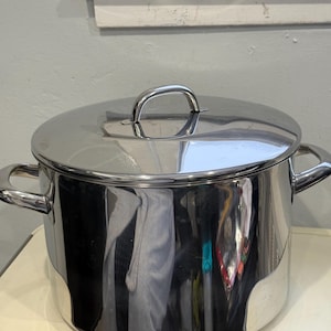 May include: A stainless steel stockpot with a matching lid. The pot has two side handles and a loop handle on the lid. The pot is sitting on a white surface.