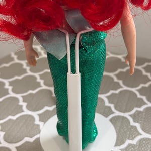 May include: A doll with bright red hair and a green sequined mermaid tail. The doll is supported by a white stand. A light blue bow is visible at the waist. The doll's arms are visible, and the skin tone is fair.