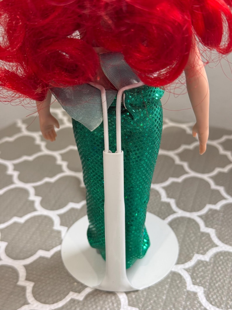 May include: A doll with bright red hair and a green sequined mermaid tail. The doll is supported by a white stand. A light blue bow is visible at the waist. The doll's arms are visible, and the skin tone is fair.